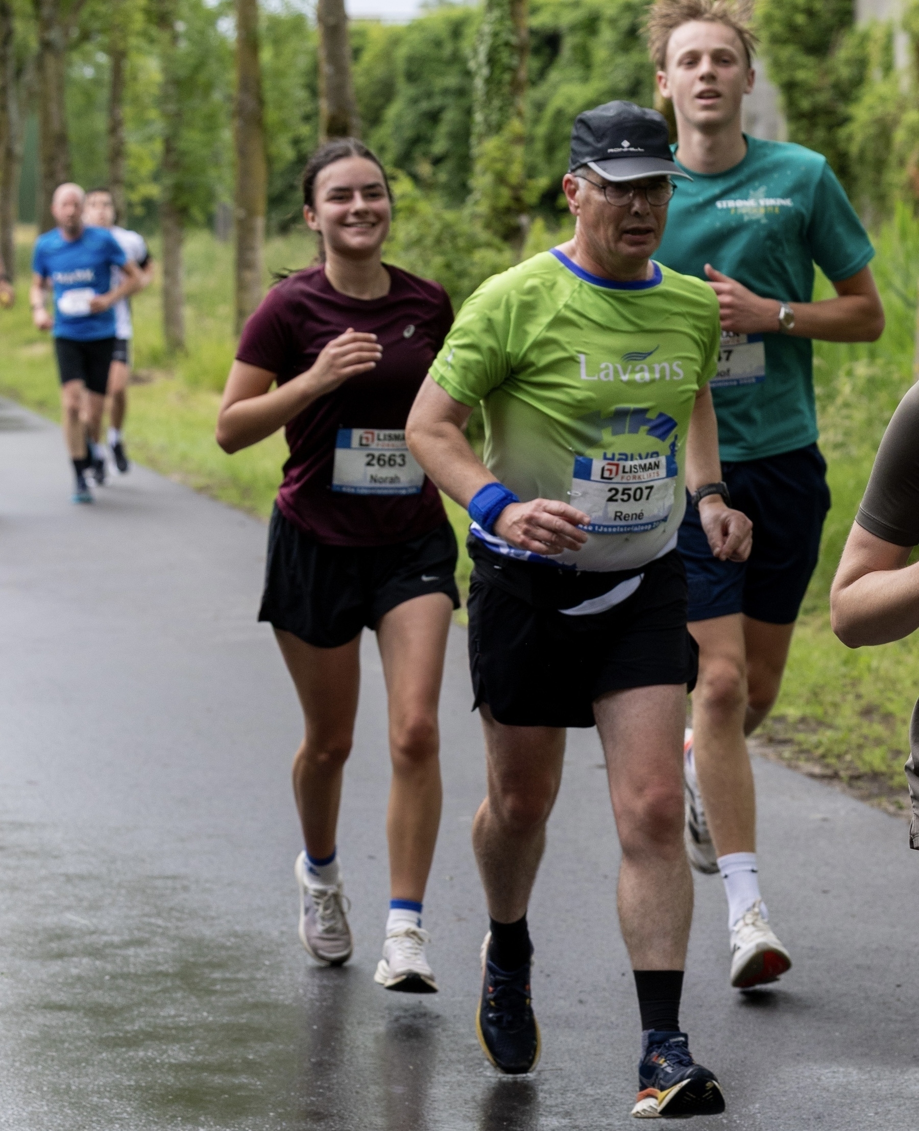 A group of runners, with race numbers pinned to their shirts, are participating in an outdoor event on a tree-lined path.